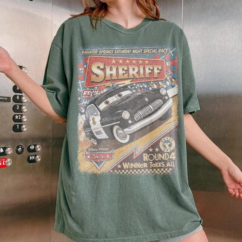May include: A sage green short-sleeve t-shirt featuring a graphic print of the Sheriff character from the movie Cars. The design includes the text Radiator Springs Saturday Night Special Race, Sheriff, Round 4, and Winner Takes All with stars.