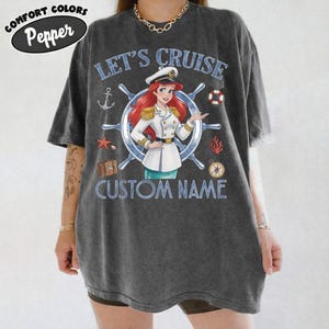Pu&ograve; includere: T-shirt Comfort Colors grigio scuro con un personaggio dei cartoni animati in uniforme da capitano, la scritta "LET'S CRUISE" e "CUSTOM NAME". Il design include elementi nautici come un'ancora, una stella marina e un timone. La maglietta ha la scritta "Pepper".
