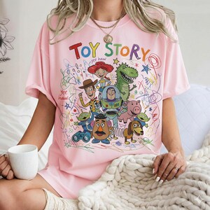 May include: A light pink t-shirt featuring the words "TOY STORY" in colorful letters, along with cartoon characters like Woody, Buzz Lightyear, and Rex. The design includes scribbled lines and stars, creating a playful aesthetic.