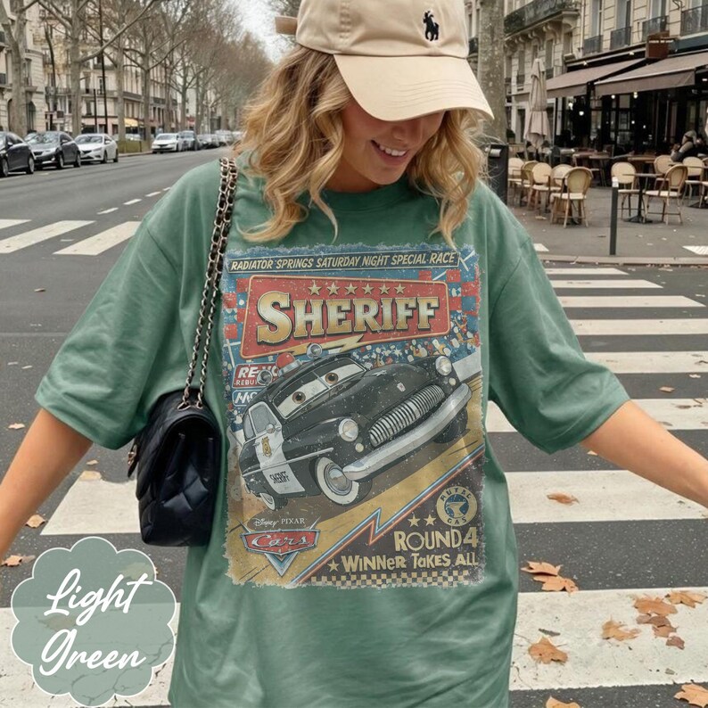May include: A sage green t-shirt featuring a graphic print of the character Sheriff from the movie Cars. The design includes the text Radiator Springs Saturday Night Special Race, Sheriff, and Round 4 Winner Takes All, with a black police car illustration.