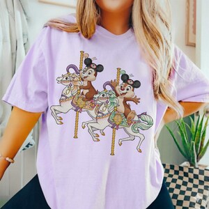 May include: A light purple t-shirt featuring a carousel design with two cartoon characters riding white horses. The carousel has gold poles and the characters are wearing hats. The shirt is made of a soft material.