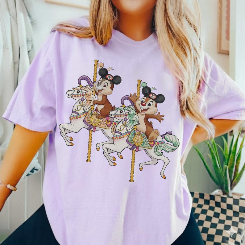 May include: A light purple t-shirt featuring a carousel design with two cartoon characters riding white horses. The carousel has gold poles and the characters are wearing hats. The shirt is made of a soft material.