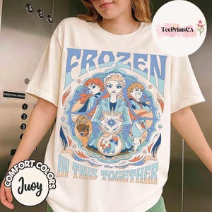 K&ouml;nnte beinhalten: Cremefarbenes T-Shirt mit einer farbenfrohen Grafik aus Disneys Frozen. Das Design enth&auml;lt die W&ouml;rter "FROZEN" und "IN THIS TOGETHER" mit Bildern von Elsa, Anna, Kristoff, Olaf und Sven. Das Shirt hat kurze &Auml;rmel und einen Rundhalsausschnitt.