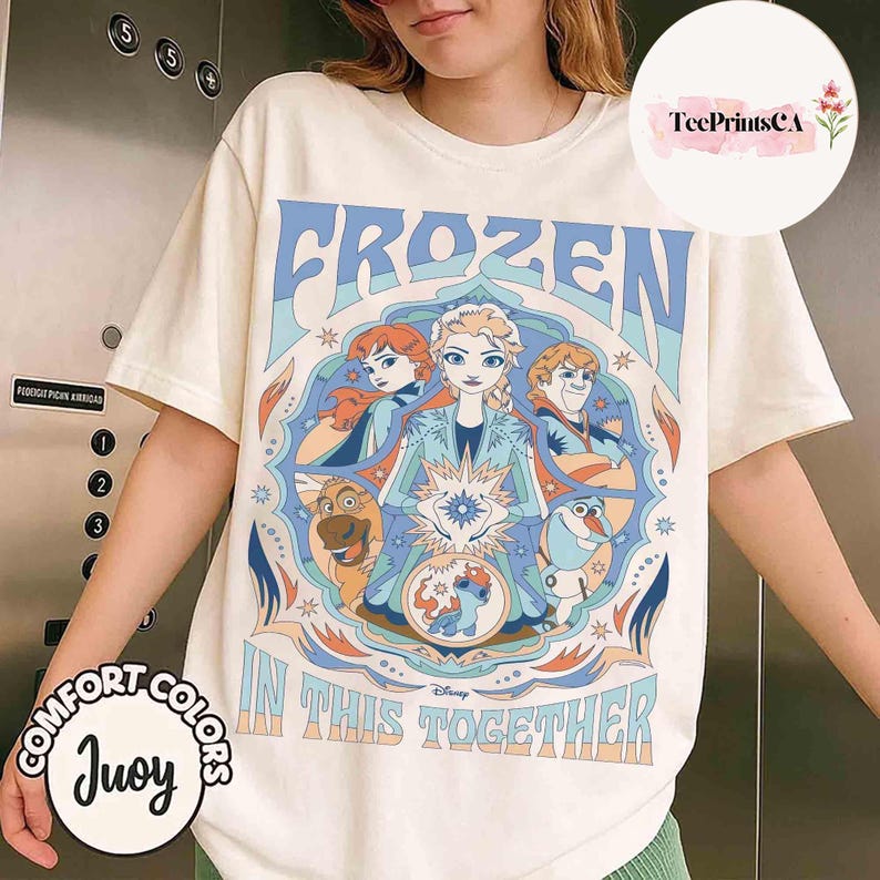 K&ouml;nnte beinhalten: Cremefarbenes T-Shirt mit einer farbenfrohen Grafik aus Disneys Frozen. Das Design enth&auml;lt die W&ouml;rter "FROZEN" und "IN THIS TOGETHER" mit Bildern von Elsa, Anna, Kristoff, Olaf und Sven. Das Shirt hat kurze &Auml;rmel und einen Rundhalsausschnitt.