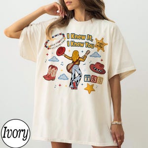 May include: An ivory t-shirt featuring a graphic print of a person with long blonde hair playing a guitar, surrounded by cowboy boots, hats, clouds, stars, and alphabet blocks. The text reads I Knew It, I Knew You, Infinity and Beyond, and You've got a friend in me.