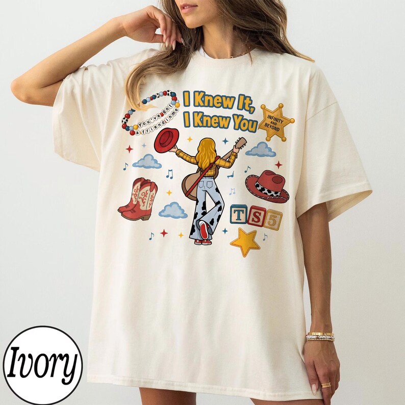 May include: An ivory t-shirt featuring a graphic print of a person with long blonde hair playing a guitar, surrounded by cowboy boots, hats, clouds, stars, and alphabet blocks. The text reads I Knew It, I Knew You, Infinity and Beyond, and You've got a friend in me.