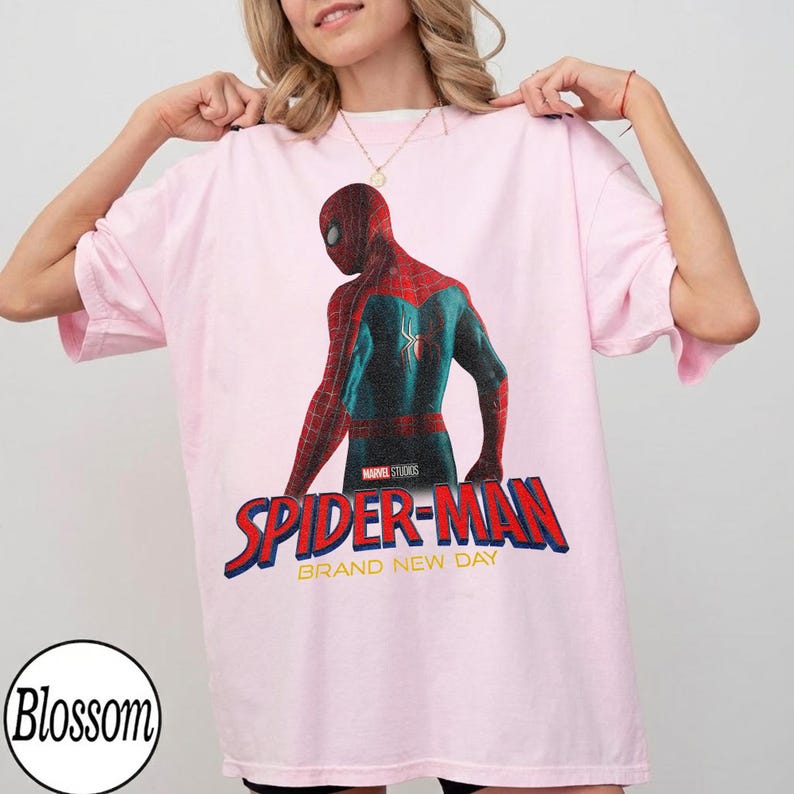 May include: Light pink t-shirt featuring a graphic of Spider-Man in a red and blue suit, with the text "SPIDER-MAN" in red and blue, and "BRAND NEW DAY" in white. The Marvel Studios logo is also visible.