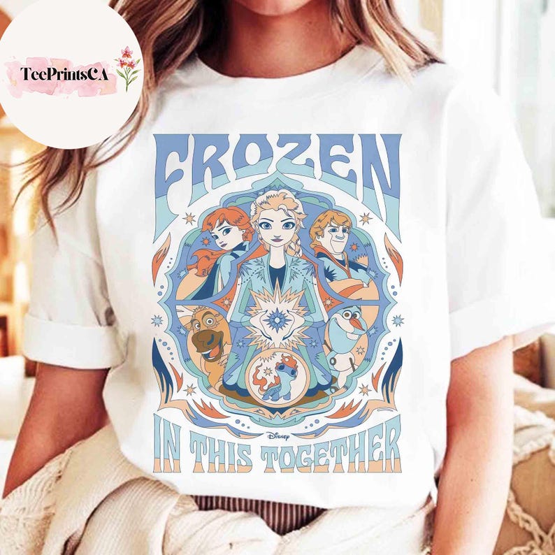 K&ouml;nnte beinhalten: Wei&szlig;es T-Shirt mit einem farbenfrohen Grafikdesign aus dem Disney-Film "Frozen". Das Design enth&auml;lt das Wort "FROZEN" in Blau, zusammen mit Bildern von Charakteren und dem Satz "IN THIS TOGETHER".