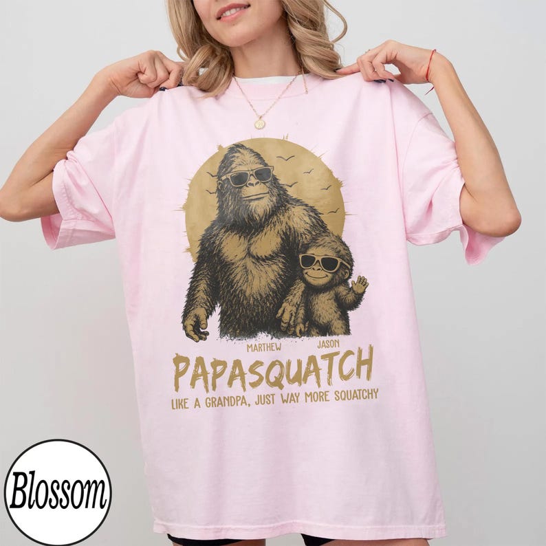 May include: A light pink t-shirt featuring a graphic of a Bigfoot with a child Bigfoot, both wearing sunglasses. The text reads "PAPASQUATCH" and "LIKE A GRANDPA, JUST WAY MORE SQUATCHY."