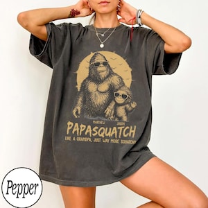 May include: Dark gray t-shirt featuring a Bigfoot graphic wearing sunglasses with a child Bigfoot. The text reads "PAPASQUATCH" and "LIKE A GRANDPA, JUST WAY MORE SQUATCHY". A fun, novelty tee.