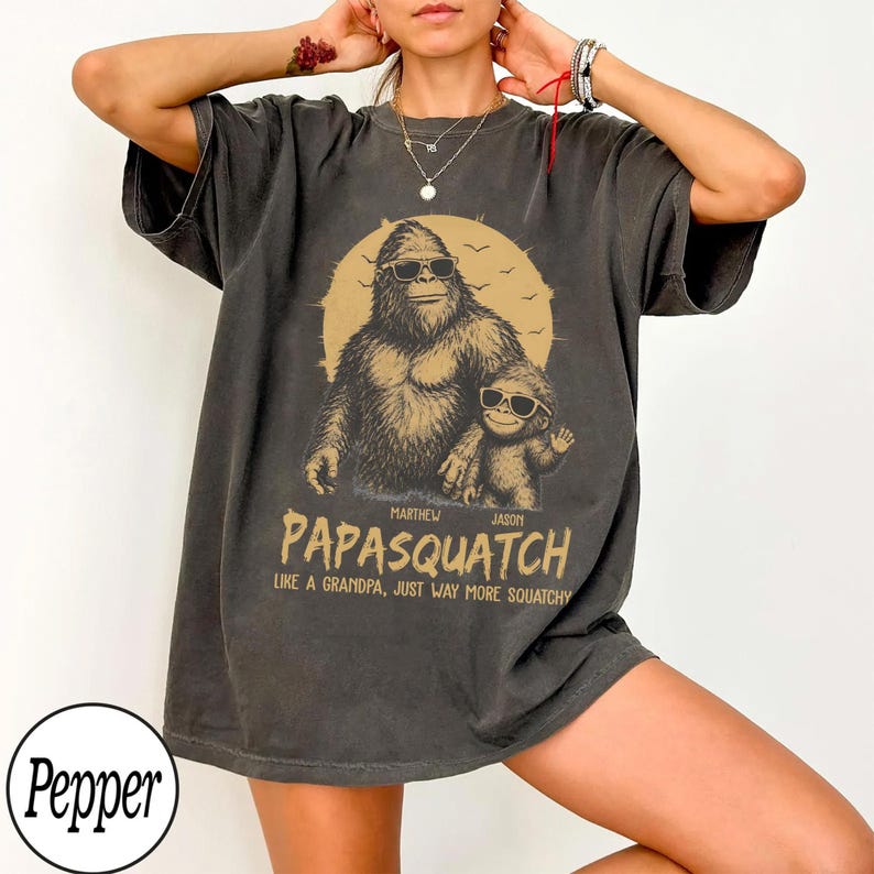 May include: Dark gray t-shirt featuring a Bigfoot graphic wearing sunglasses with a child Bigfoot. The text reads "PAPASQUATCH" and "LIKE A GRANDPA, JUST WAY MORE SQUATCHY". A fun, novelty tee.