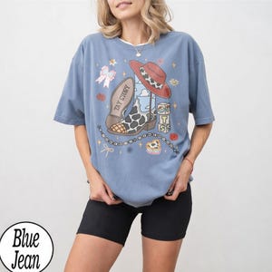 May include: A dusty blue short-sleeve t-shirt featuring a graphic print of a cow-print cowboy boot, a red hat, alphabet blocks spelling TS, and the text TAY STORY. The design includes small stars, a bow, and a beaded string graphic element.