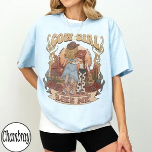 Może przedstawiać: Jasnoniebieski t-shirt z nadrukiem graficznym przedstawiającym dwie postacie w kowbojskich kapeluszach na tle pustynnego krajobrazu z kaktusami i r&oacute;żami. Napis głosi COW GIRL LIKE ME. Koszulka ma kr&oacute;tkie rękawy i okrągły dekolt o luźnym kroju.