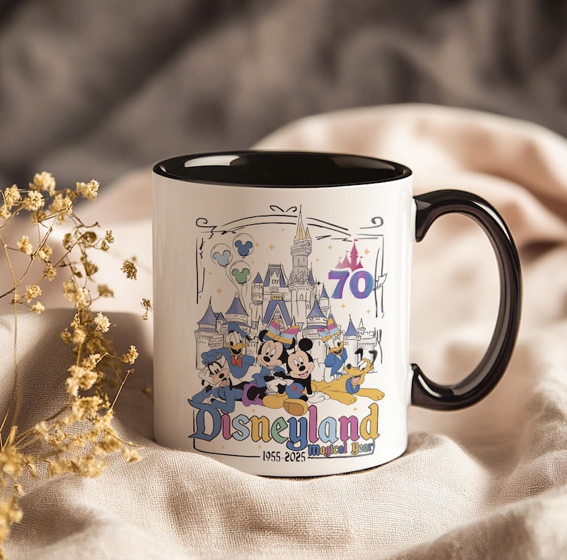 May include: White ceramic mug with a black interior and handle. The mug features a colorful illustration of Disneyland characters and the castle, with the text "Disneyland" and "70". The mug also has the text "1955-2025 Magical Years".