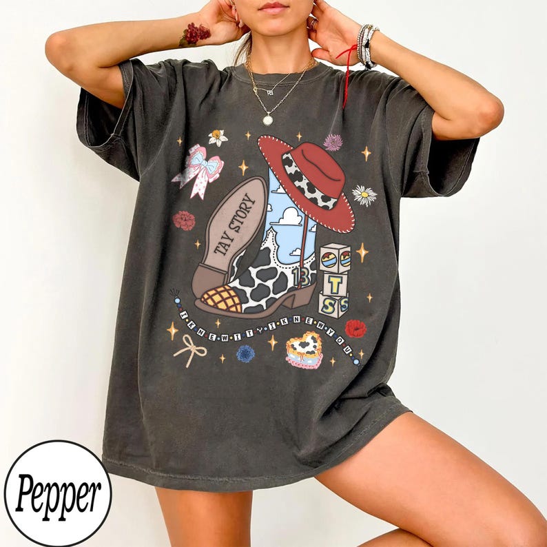 May include: A charcoal gray short-sleeve t-shirt featuring a graphic print of a cow-print cowboy boot, a red hat, and blocks with the letters T, S, and 13. The text TAY STORY is on the boot, and the text BE-NEW-WIT-TH-KN-EN-YOU is printed below.