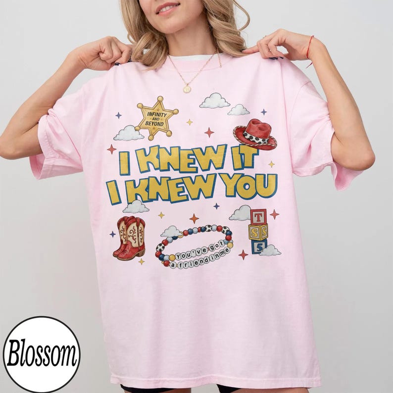May include: A light pink t-shirt featuring a graphic print with the text I KNEW IT I KNEW YOU, a gold sheriff star badge labeled INFINITY AND BEYOND, a red cowboy hat, brown cowboy boots, a beaded bracelet with the text YOU'VE GOT A FRIEND IN ME, and alphabet blocks.