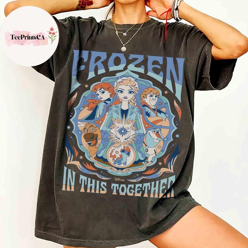 K&ouml;nnte beinhalten: Dunkelgraues T-Shirt mit einer farbenfrohen Grafik aus dem Disney-Film "Die Eisk&ouml;nigin". Das Design enth&auml;lt das Wort "FROZEN" in Blau und den Satz "IN THIS TOGETHER". Die Grafik zeigt Elsa, Anna, Kristoff, Olaf, Sven und Stitch.