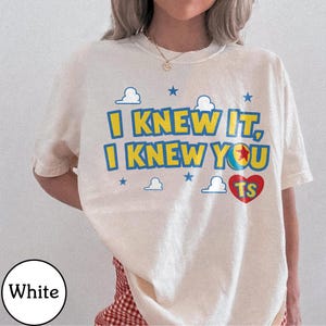 Pu&ograve; includere: Una t-shirt bianca a maniche corte con il testo giallo e blu I KNEW IT, I KNEW YOU, una grafica a palla colorata, un cuore rosso contenente le lettere TS e piccole stelle blu e icone di nuvole bianche stampate sul petto.