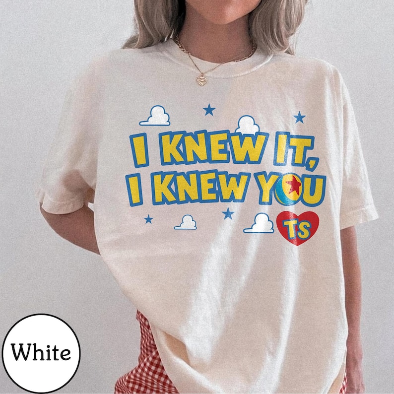 Pu&ograve; includere: Una t-shirt bianca a maniche corte con il testo giallo e blu I KNEW IT, I KNEW YOU, una grafica a palla colorata, un cuore rosso contenente le lettere TS e piccole stelle blu e icone di nuvole bianche stampate sul petto.