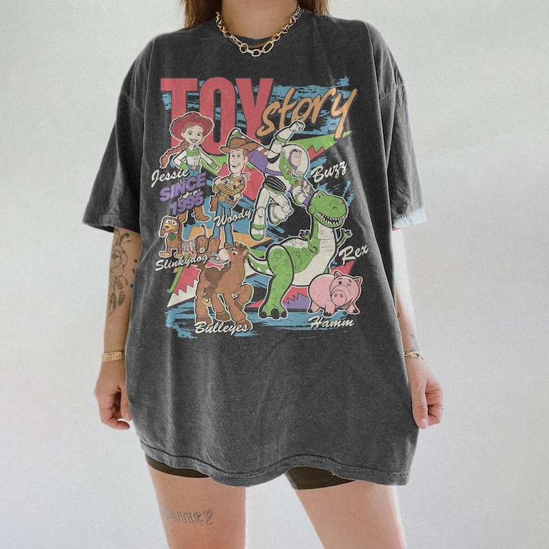Puede incluir: Camiseta gris oscuro con el texto "Toy Story" en una fuente retro colorida. La camiseta muestra personajes de dibujos animados como Woody, Buzz Lightyear y Jessie. Tambi&eacute;n es visible el texto "Since 1995".