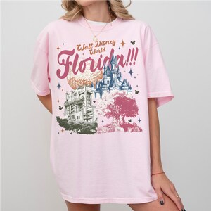 May include: Light pink t-shirt featuring a graphic design of Walt Disney World Florida. The design includes the words "Walt Disney World Florida!!!" in a cursive font, along with illustrations of the Spaceship Earth, Cinderella Castle, and the Hollywood Tower Hotel.