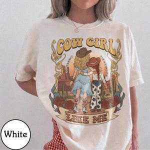 Może przedstawiać: Biały t-shirt z nadrukiem graficznym przedstawiającym dwie postacie w kowbojskich kapeluszach na tle pustynnego krajobrazu z kaktusami i r&oacute;żami. Tekst głosi COW GIRL LIKE ME oraz I KNEW YOU, w otoczeniu ozdobnych złotych ornament&oacute;w.