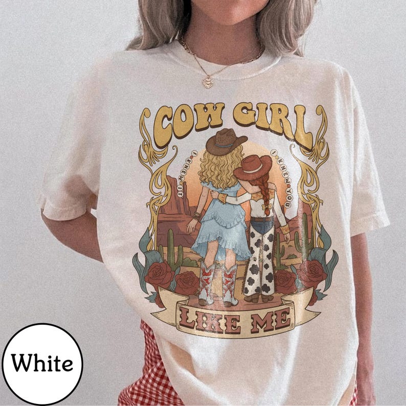 Może przedstawiać: Biały t-shirt z nadrukiem graficznym przedstawiającym dwie postacie w kowbojskich kapeluszach na tle pustynnego krajobrazu z kaktusami i r&oacute;żami. Tekst głosi COW GIRL LIKE ME oraz I KNEW YOU, w otoczeniu ozdobnych złotych ornament&oacute;w.
