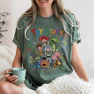 May include: Green t-shirt featuring the words "TOY STORY" in colorful letters above a graphic of characters from the animated film. The design includes Woody, Buzz Lightyear, and other recognizable characters.