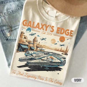 Puede incluir: Camiseta de color marfil con un gráfico del Halcón Milenario y el texto "GALAXY'S EDGE Est. 2019." El diseño incluye ilustraciones de edificios, naves espaciales y la frase "The Most Legendary Place In The Galaxy."
