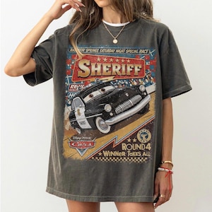 May include: A charcoal gray short-sleeve t-shirt featuring a graphic print of the character Sheriff from Disney Pixar Cars. The design includes the text Radiator Springs Saturday Night Special Race, Sheriff, and Round 4 Winner Takes All.