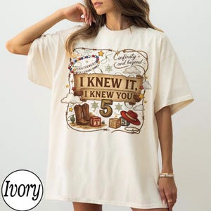 May include: An ivory t-shirt featuring a graphic print with a brown banner that reads I KNEW IT, I KNEW YOU, the number 5, and the text infinity and beyond. The design includes cowboy boots, a red hat, alphabet blocks, and a beaded bracelet.