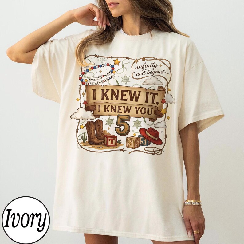 May include: An ivory t-shirt featuring a graphic print with a brown banner that reads I KNEW IT, I KNEW YOU, the number 5, and the text infinity and beyond. The design includes cowboy boots, a red hat, alphabet blocks, and a beaded bracelet.