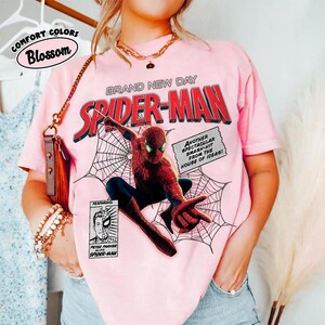 Op de afbeelding: Zachtroze T-shirt met een Spider-Man-afbeelding. Het ontwerp bevat de woorden "BRAND NEW DAY SPIDER-MAN" in rood en wit. Extra tekst luidt "ANOTHER SPECTACULAR SMASH-HIT FROM THE HOUSE OF IDEAS!" en "FEATURING PETER PARKER ALIAS SPIDER-MAN."