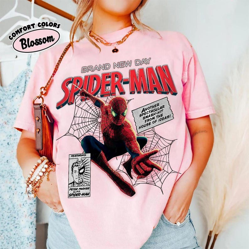 Op de afbeelding: Zachtroze T-shirt met een Spider-Man-afbeelding. Het ontwerp bevat de woorden "BRAND NEW DAY SPIDER-MAN" in rood en wit. Extra tekst luidt "ANOTHER SPECTACULAR SMASH-HIT FROM THE HOUSE OF IDEAS!" en "FEATURING PETER PARKER ALIAS SPIDER-MAN."