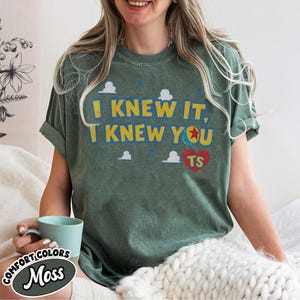 Pu&ograve; includere: Una t-shirt a maniche corte color verde muschio con il testo giallo I KNEW IT, I KNEW YOU, una grafica a palla colorata che sostituisce la lettera O, piccole icone di nuvole bianche e un cuore rosso con le lettere TS stampate sul petto.