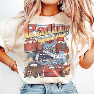 May include: Cream-colored sweatshirt featuring a graphic of various cartoon cars with the text "Radiator Springs SINCE 2012". The design includes a fire truck, police car, and race car.
