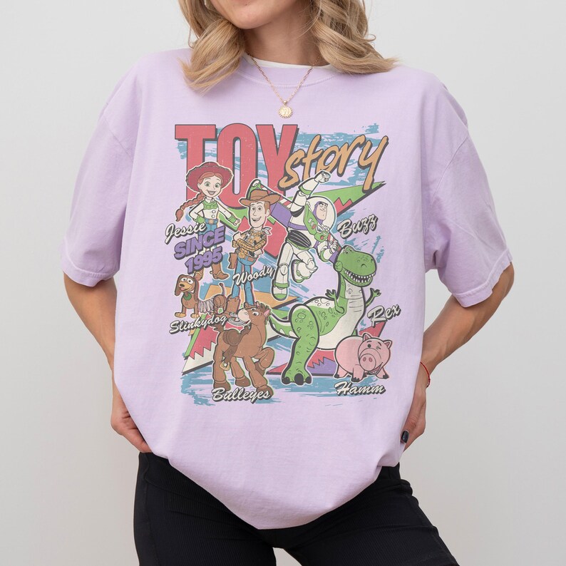 Puede incluir: Camiseta lila claro con un dise&ntilde;o de Toy Story. El dise&ntilde;o incluye personajes como Woody, Buzz Lightyear y Rex, con el texto "TOY STORY" en la parte superior. Debajo est&aacute;n los nombres de los personajes y "Jessie Since 1995". El estilo es retro y vintage.