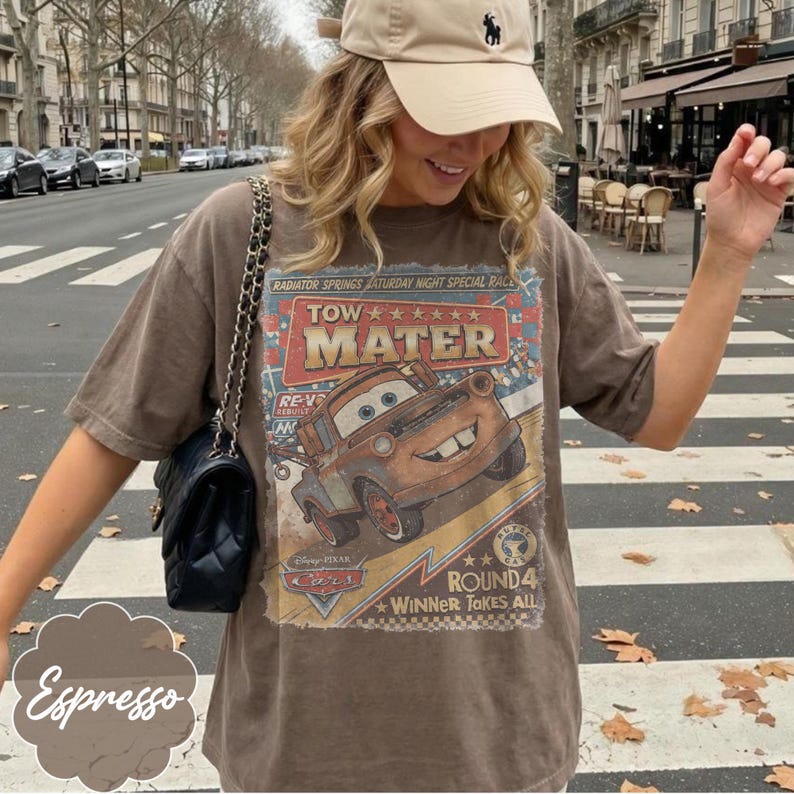 May include: A brown short-sleeve t-shirt featuring a graphic print of the character Tow Mater from the Disney Pixar Cars movie. The print includes the text Tow Mater, Radiator Springs Saturday Night Special Race, and Winner Takes All.