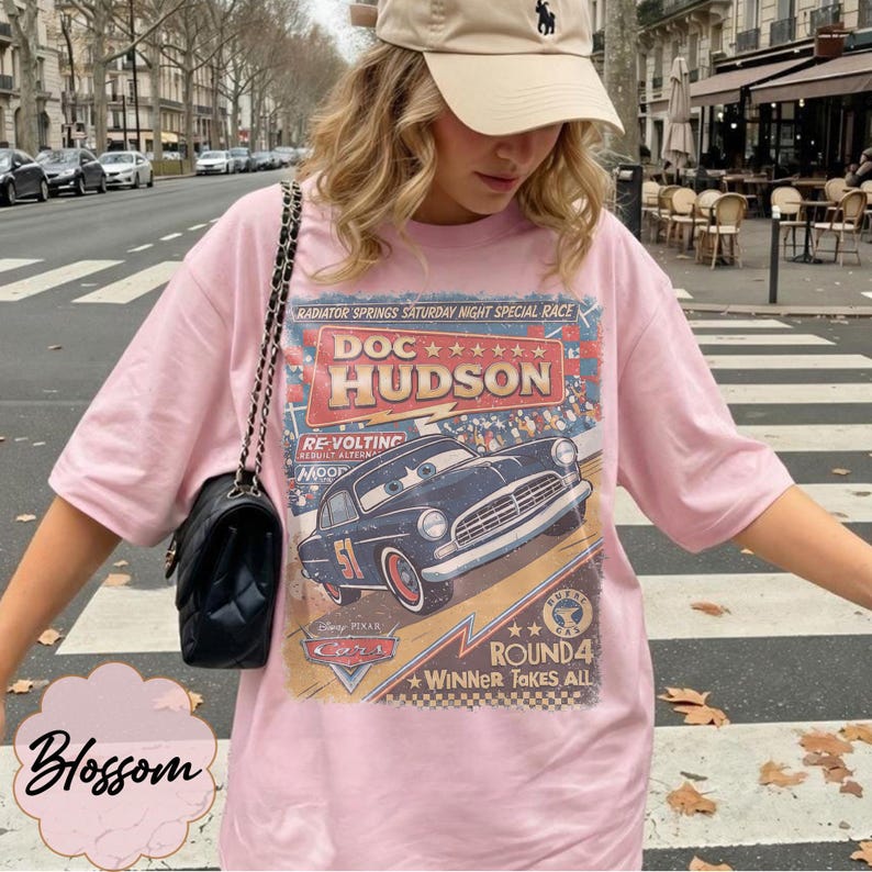 May include: A light pink t-shirt featuring a graphic print of Doc Hudson from the movie Cars. The design includes the text Radiator Springs Saturday Night Special Race, Doc Hudson, Re-Volting, Round 4 Winner Takes All, and the Disney Pixar Cars logo.