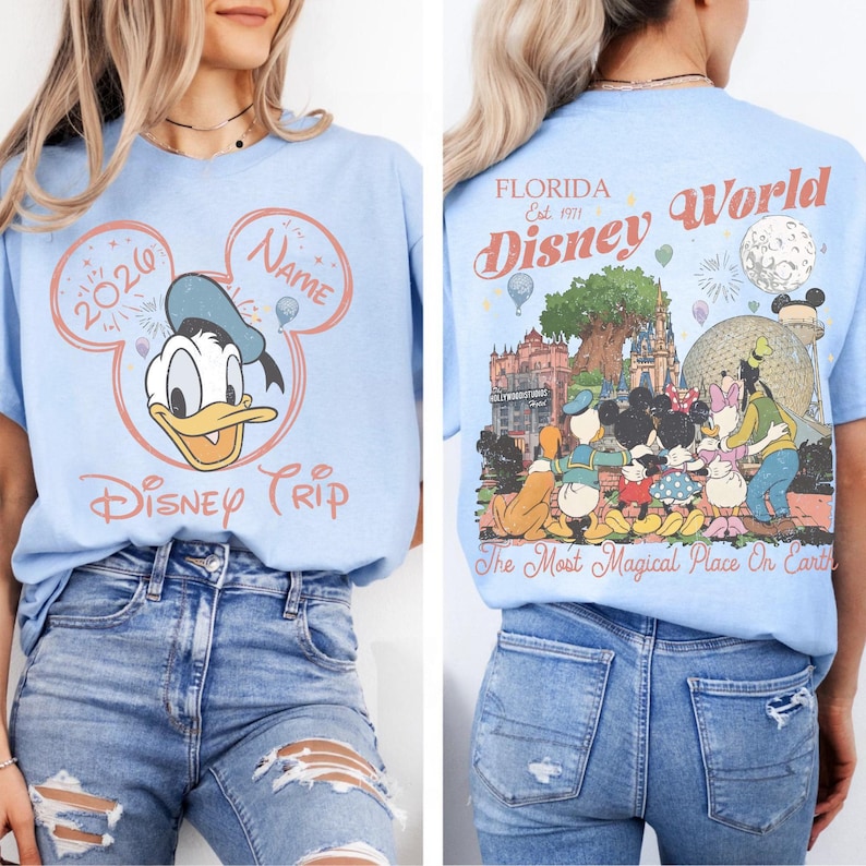 K&ouml;nnte beinhalten: Hellblaues T-Shirt mit Donald Duck und den Aufschriften "2026" und "Name" in einem Mickey-Mouse-Ohren-Design. Auf der R&uuml;ckseite des T-Shirts befindet sich eine Disney World-Grafik mit dem Text "Florida Disney World" und "The Most Magical Place On Earth."