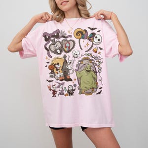 May include: A light pink t-shirt featuring a graphic print of various characters and icons from The Nightmare Before Christmas, including Jack Skellington, Sally, Oogie Boogie, Zero, and Lock, Shock, and Barrel, arranged in a collage style.