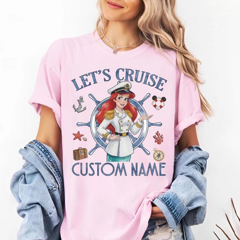 Pu&ograve; includere: T-shirt rosa chiaro con le scritte blu "LET'S CRUISE" e "CUSTOM NAME". La grafica presenta un cartone animato di una donna in uniforme da capitano, un timone e elementi nautici.