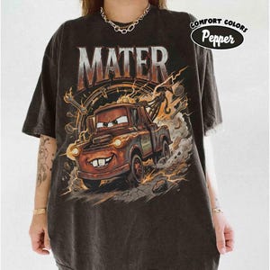 May include: Dark gray t-shirt featuring a graphic of Mater from the movie Cars. The design includes the word "MATER" in large, distressed lettering above the character. The shirt has a distressed look and the words "COMFORT COLORS Pepper" are printed on the upper right.