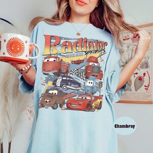 May include: Light blue t-shirt featuring a graphic of various cartoon cars with the text "Radiator Springs Since 2012". The shirt also has the word "Chambray" printed on it. A red and white mug with a sun design is held in the image.