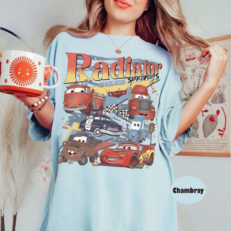 May include: Light blue t-shirt featuring a graphic of various cartoon cars with the text "Radiator Springs Since 2012". The shirt also has the word "Chambray" printed on it. A red and white mug with a sun design is held in the image.