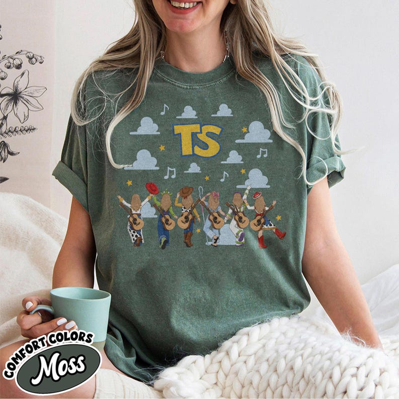 May include: A moss green short-sleeve t-shirt featuring a graphic print of six figures playing acoustic guitars. The design includes the yellow text TS, light blue clouds, musical notes, and small yellow stars scattered across the front.