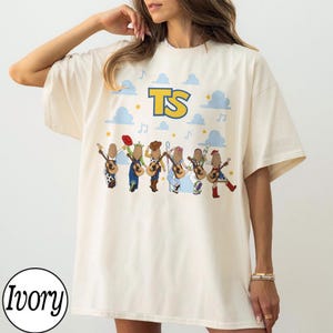 May include: An ivory short-sleeve t-shirt featuring a graphic print of six figures playing acoustic guitars. Above the figures, the letters TS are printed in yellow with a blue outline, surrounded by light blue clouds, stars, and musical notes.