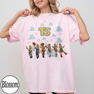 May include: A light pink t-shirt featuring the text TS in yellow with a blue outline. Below the text are six illustrated figures wearing various outfits and holding acoustic guitars, surrounded by light blue clouds, yellow stars, and musical notes.