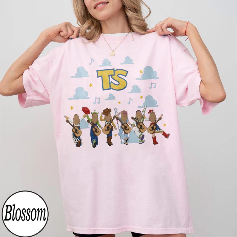 May include: A light pink t-shirt featuring the text TS in yellow with a blue outline. Below the text are six illustrated figures wearing various outfits and holding acoustic guitars, surrounded by light blue clouds, yellow stars, and musical notes.