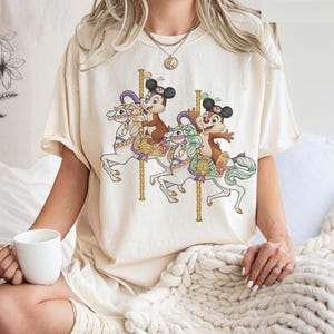 May include: Cream-colored t-shirt featuring a carousel scene with cartoon characters. The design shows two chipmunk-like characters riding white carousel horses with gold poles. The horses have colorful saddles and manes. The shirt has short sleeves.